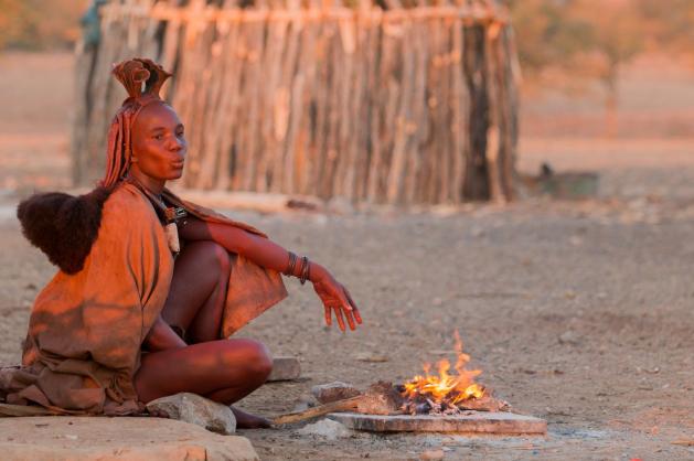 Žena z kmene Himba sedící u ohně před tradiční chýší.